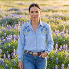 Sky blue chambray pearl-snap Cowgirl+ Blouse with bluebonnet embroidery on the yoke and cuffs, tucked into jeans with a concho belt