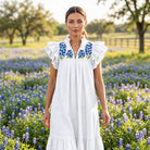 White denim midi dress with blue bluebonnet embroidery on the yoke, ruffle collar and ruffle sleeves, worn in a Texas bluebonnet field